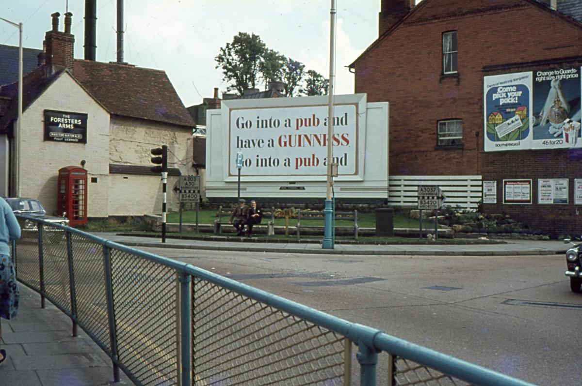 The junction of Winchester Street and London Street. The site of advertisement hoarding is now built over. The blue fence and rail was to protect pedestrians from the A303 which ran from Bridge Street up London Street. From the Jeffery Saunders collection